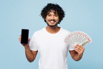 Young happy Indian man wear white t-shirt casual clothes hold fan of cash money in dollar banknotes use blank screen mobile cell phone isolated on plain pastel blue cyan background. Lifestyle concept.