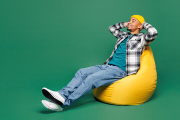 Wall Mural - Full body young man of African American ethnicity he wear shirt blue t-shirt yellow hat sit in bag chair hold hands behind neck isolated on plain green background studio portrait. Lifestyle concept.