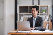 © wichayada - Young Asian man working on laptop in modern office analyze accounting, report real estate investment information.
