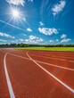   The sun shines brightly over a grassy area where a running track cuts through, surrounded by a blue sky dotted with white, wispy clouds