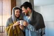© NDABCREATIVITY - Happy young couple with hot drinks cuddling under warm blanket at home
