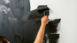 Close-up view of a person's hand using a roller to paint a wall black, showing rich texture and motion.
