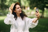 Fototapeta Niebo - Young beautiful girl in a hat makes a selfie on her phone in the park. High quality photo