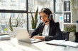 © forenna - smart young businesswoman is captured wearing headphones, fully immersed in a language learning webinar on her laptop. With pen in hand, she diligently takes notes during online co