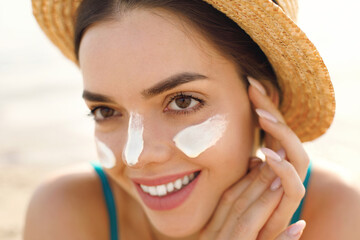 Beautiful young woman at beach applying sunscreen on face and looking at camera. Beauty girl applying suntan lotion at sea. Portrait of happy woman with healthy skin applying sunblock on cheek.