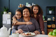 Front view portrait of happy African American grandmother with two girls embracing at kitchen table and looking at camera