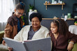 Portrait of smiling senior African American woman with two children looking at family photo album together at home