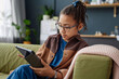Portrait of young African American girl using digital tablet while sitting on couch at home and browsing internet copy space