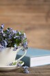 Beautiful forget-me-not flowers in cup, saucer and book on wooden table, closeup. Space for text