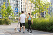 © CinemaF - A young athletic couple walks outdoors with pet dog on a warm summer day on the street in a park town surrounded by colorful modern residential buildings. The concept of walking as a complete workout