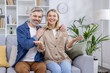 © Liubomir - An attractive, mature couple sitting comfortably on a sofa at home, presenting a relaxed and welcoming smile to the viewers. The setting is bright and cozy, decorated with plants and modern