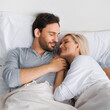 Portrait of young beautiful couple looking at each other, lay in home bed. Blond woman, brunette bearded man in love, relationship, happy family, waiting child concept. Square image.