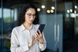 © Liubomir - A focused Hispanic businesswoman in glasses uses her smartphone, possibly reading an important message or checking business updates in a modern office setting.