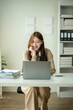 © apichat - A woman is sitting at a desk with a laptop and smiling. She is wearing a brown shirt and tan pants. The room has a white wall and a bookshelf with many books