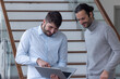 © Tof - Photographie - deux jeune hommes d'affaire ou employés de bureau discutent debout dans les locaux de leur entreprise. l'un d'eux porte un ordinateur portable