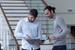 © Tof - Photographie - deux jeune hommes d'affaire ou employés de bureau discutent debout dans les locaux de leur entreprise. l'un d'eux porte un ordinateur portable