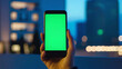 hand holding smartphone with blank green screen against a cityscape. indoor setting and technological elements. Ideal for themes related to app development, smart home technology, or urban living