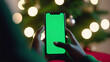 hand holding a smartphone with blank green screen against a festive Christmas lights background. Displaying mobile apps for holiday shopping or celebration experience
