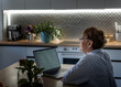 Senior pensioner woman using laptop while sitting at home in the kitchen in the evening