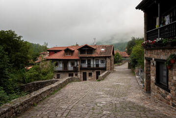 Wall Mural - An old small village with cobblestone streets and traditional houses with red tiles roofs with foggy mountains in the background. Barcena Mayor, Saja-Besaya Natural Park, Cantabria, Spain