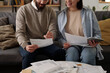 Cropped shot of young couple in casualwear sitting on couch and looking through text of financial document held by bearded man