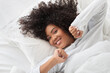 © Prostock-studio - A young Hispanic woman with curly hair is lying in bed, exhibiting a relaxed and happy expression as she gently wakes up.