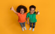 © Prostock-studio - A cheerful African American boy and girl are standing on a bright yellow surface, looking upwards into the camera with joyful smiles, giving a thumbs up gesture, top view
