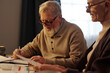 Elderly man with white hair and beard sitting by table and making notes in notepad while helping his wife with checking financial bills