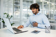 © Liubomir - A young adult male seated at his office desk shows signs of pain, clutching his stomach while looking distressed, indicating discomfort or health issues during work.