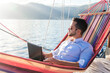 Freelancer working in traveling, using laptop, Internet. Man with computer in beach hammock. Sea summer vacation on yacht. Workplace on nature outdoors. Business trip, successful lifestyle