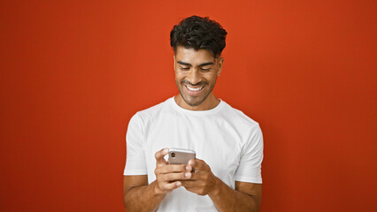 Wall Mural - Handsome young hispanic man in white shirt smiling at phone against isolated red background