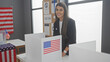 A young hispanic woman smiles at an american polling station with flags and electoral information.