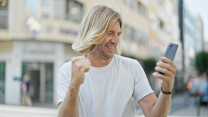 Canvas Print - Handsome young man celebrating with a fist pump while looking at his smartphone on a city street.