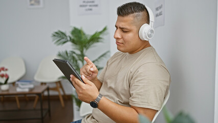 Sticker - Relaxed yet serious young latin man engrossed in his internet lifestyle, concentrating on touchpad screen and listening through headphones while sitting casual in waiting room chair