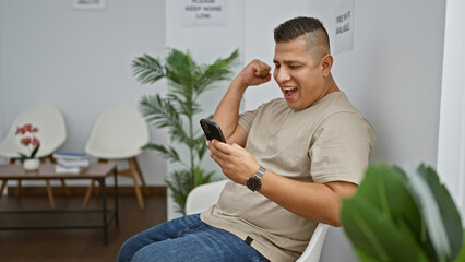 Wall Mural - Cheerful young latin man sitting in a waiting room chair, celebrating his win with a smile while using smartphone, immersed in a text conversation