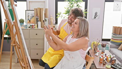 Canvas Print - Two artists, a man and woman, radiating confidence, together taking a smiling selfie in the vibrant atmosphere of their art studio