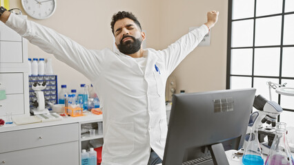 Sticker - A young hispanic man stretches in a modern laboratory setting, showing relief or a break during his work.