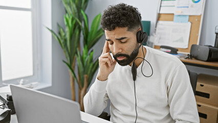 Sticker - A focused hispanic man working indoors at an office with a laptop, wearing headphones and a headset.