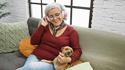 Poster - A joyful mature woman with headphones enjoys music while cuddling her dog on a cozy sofa indoors.