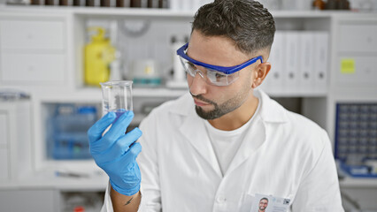 Sticker - A focused man in lab coat and safety glasses examines a test tube in a science laboratory setting.