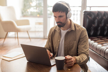 Aufkleber - A man is seated at a table, engrossed in work on his laptop. He is wearing headphones, indicating he is likely listening to something. The setting suggests a focused and productive work environment.