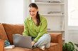 © Prostock-studio - A woman is sitting comfortably on a couch, focused on using a laptop computer. She appears engaged in her digital tasks, with a concentrated expression. The room is well-lit
