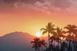 Beautiful tropical sunset with palm trees and golden sun setting over the mountains top