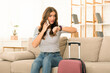 © Prostock-studio - A concerned young woman sits on the edge of a couch, gazing at her watch with a look of anxiety on her face. She is preparing to depart, evident by a suitcase beside her