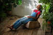 Calm black woman relaxing at home greenhouse. African American girl enjoying tranquility serenity solitude slow life resting at pouf, padded stool with laptop among green plants at indoor garden.