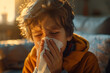 A young boy sitting on a couch is blowing his nose into a tissue, likely due to a runny nose or cold. The action conveys a common health-related scenario