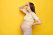 Unhappy sick unhealthy beautiful young pregnant woman wearing striped top posing isolated over yellow background suffering terrible headache touching her forehead