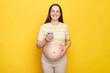 Delighted pretty happy brown haired pregnant woman with bare belly posing isolated over yellow background using mobile phone looking at camera with toothy smile