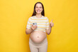 Beautiful pretty smiling Caucasian pregnant woman in casual clothing holding cell phone showing thumb up like gesture posing isolated over yellow background