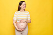 Caucasian smiling beautiful young pregnant woman wearing striped top holding her belly looking away with happy face posing isolated over yellow background